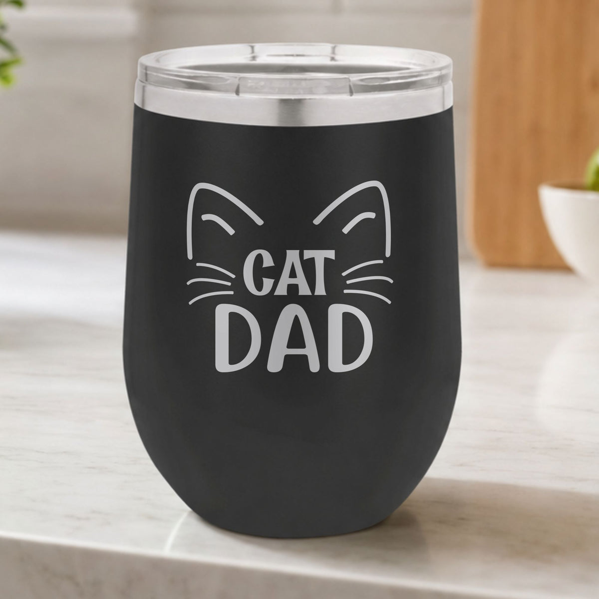 Black 12 oz stemless wine tumbler laser engraved with "Cat Dad" framed by cat ears and whiskers, on a kitchen counter