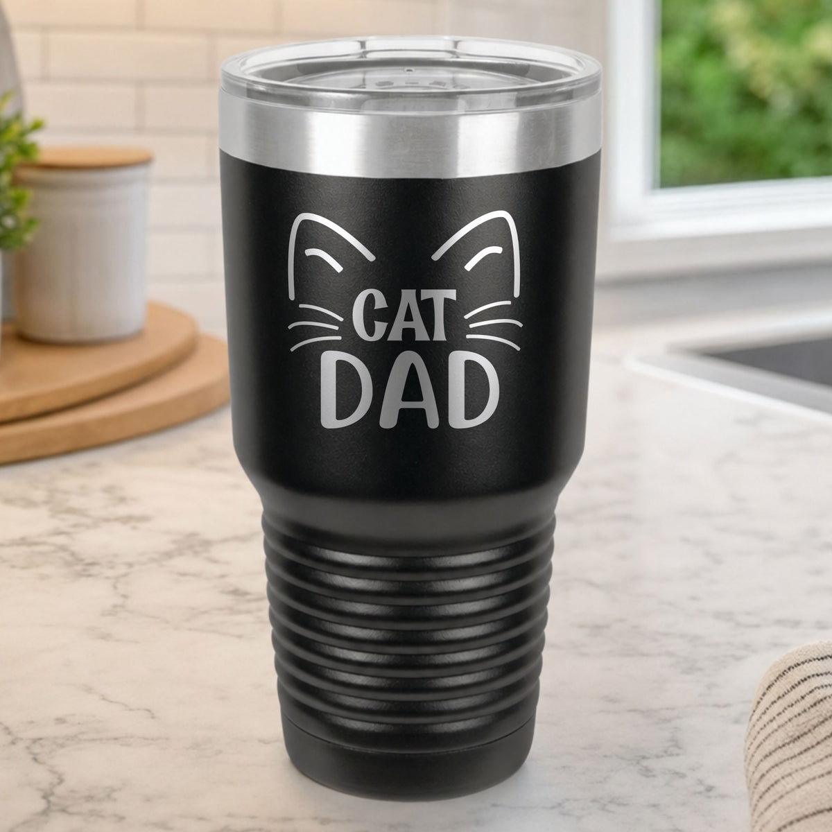 Black 30 oz tumbler laser engraved with "Cat Dad" framed by cat ears and whiskers, on a kitchen counter
