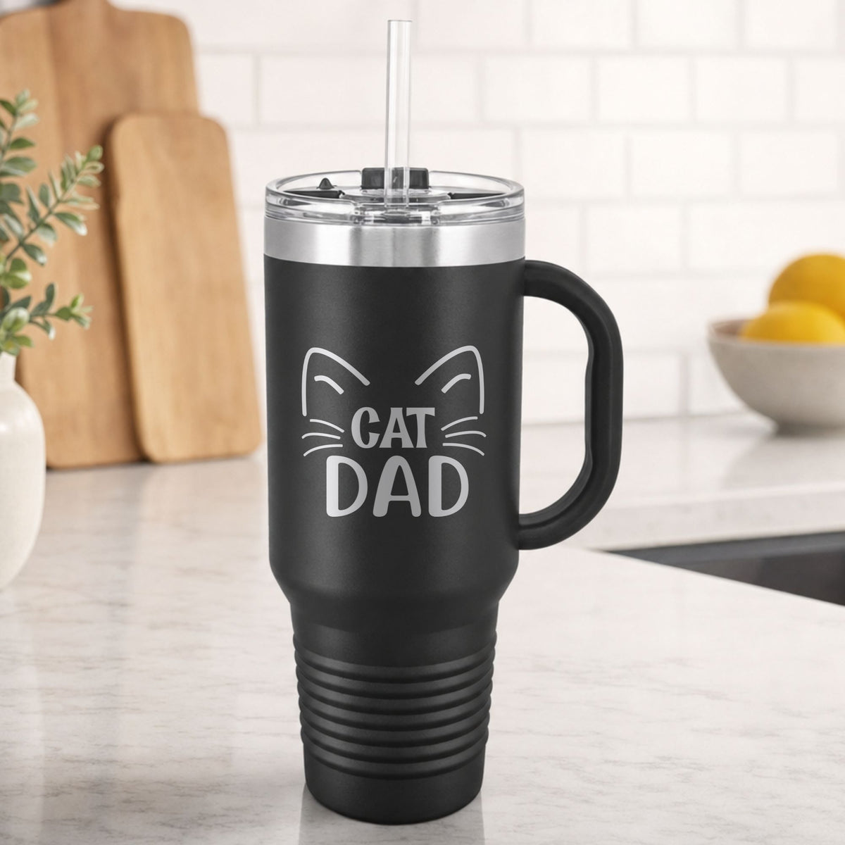 Black 40 oz tumbler with handle and straw laser engraved with "Cat Dad" framed by cat ears and whiskers, on a kitchen counter