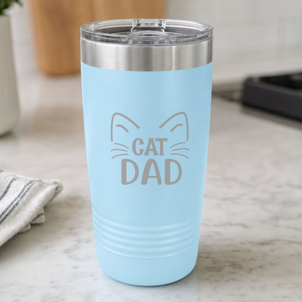 Light Blue 20 oz tumbler laser engraved with "Cat Dad" framed by cat ears and whiskers, on a kitchen counter