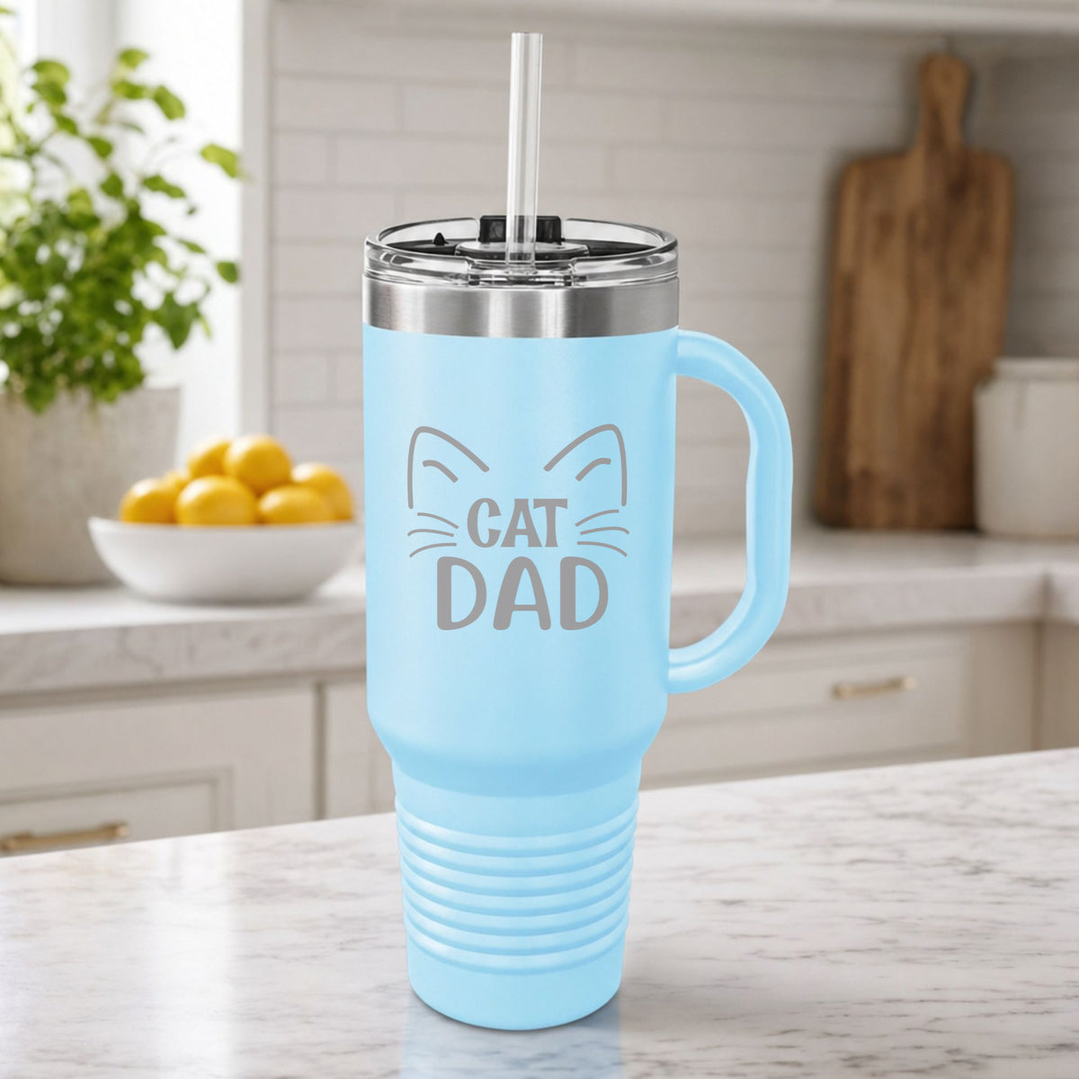 Light Blue 40 oz tumbler with handle and straw laser engraved with "Cat Dad" framed by cat ears and whiskers, on a kitchen counter