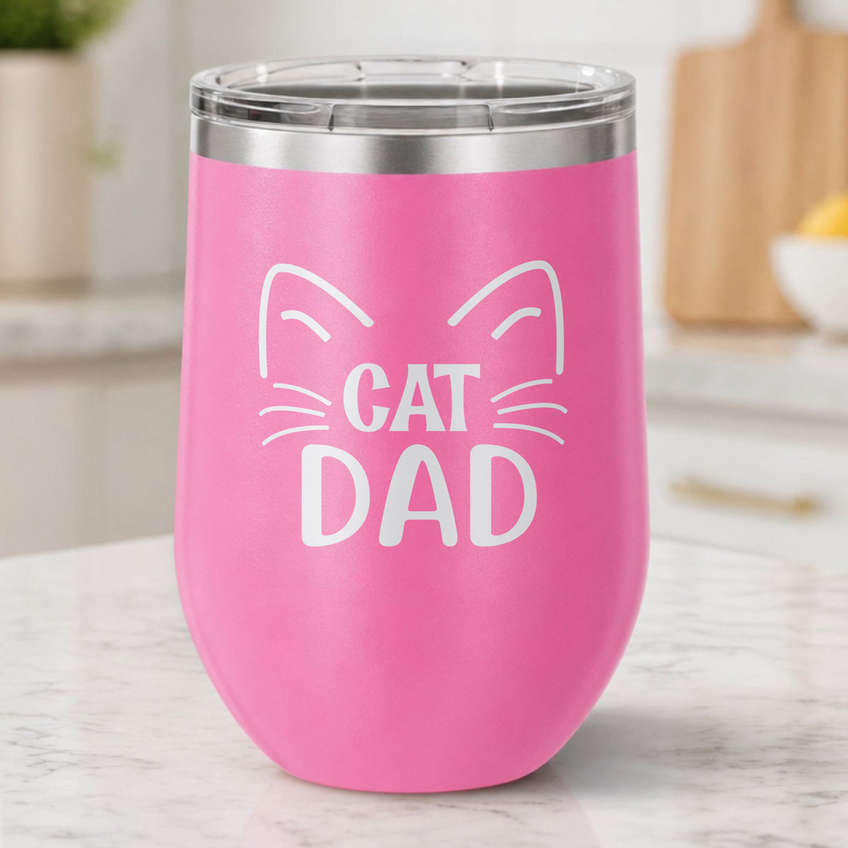 Pink 12 oz stemless wine tumbler laser engraved with "Cat Dad" framed by cat ears and whiskers, on a kitchen counter