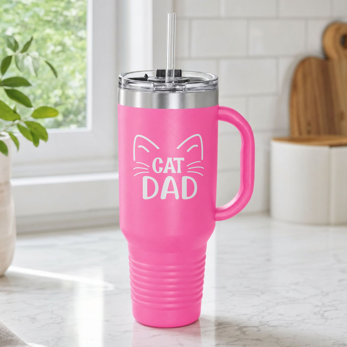 Pink 40 oz tumbler with handle and straw laser engraved with "Cat Dad" framed by cat ears and whiskers, on a kitchen counter