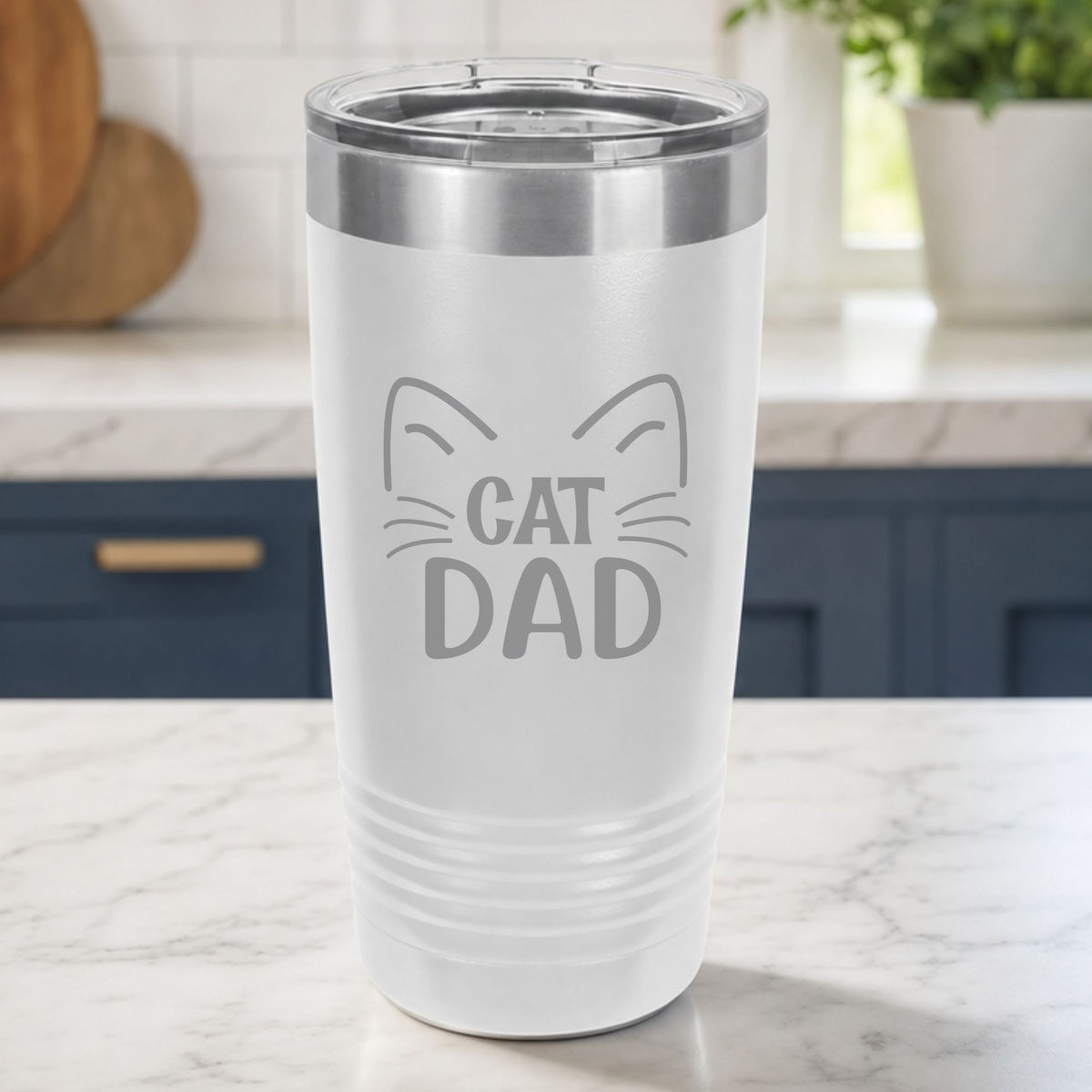 White 20 oz tumbler laser engraved with "Cat Dad" framed by cat ears and whiskers, on a kitchen counter