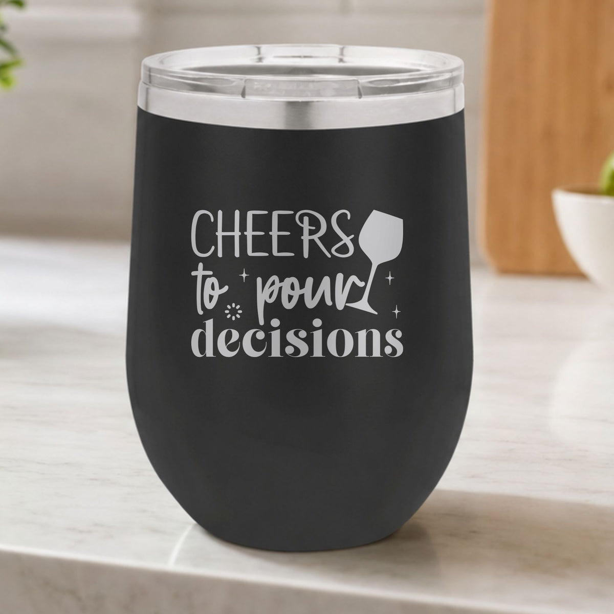 Black 12 oz stemless wine tumbler laser engraved with "Cheers to Pour Decisions", on a kitchen counter