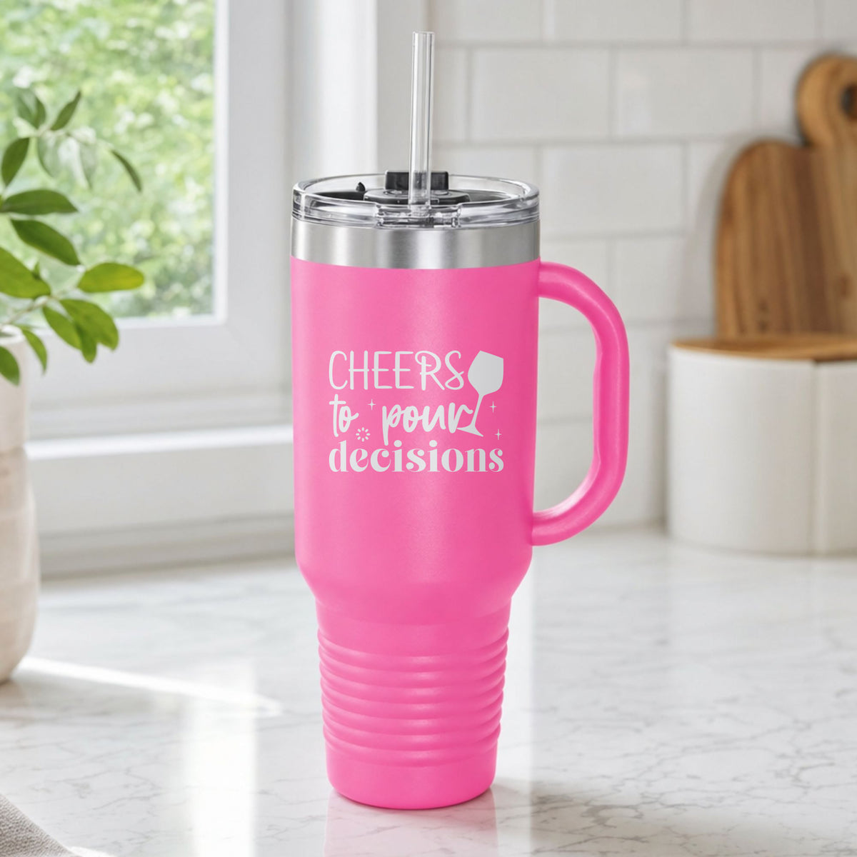 Pink 40 oz tumbler with handle and straw laser engraved with "Cheers to Pour Decisions", on a kitchen counter