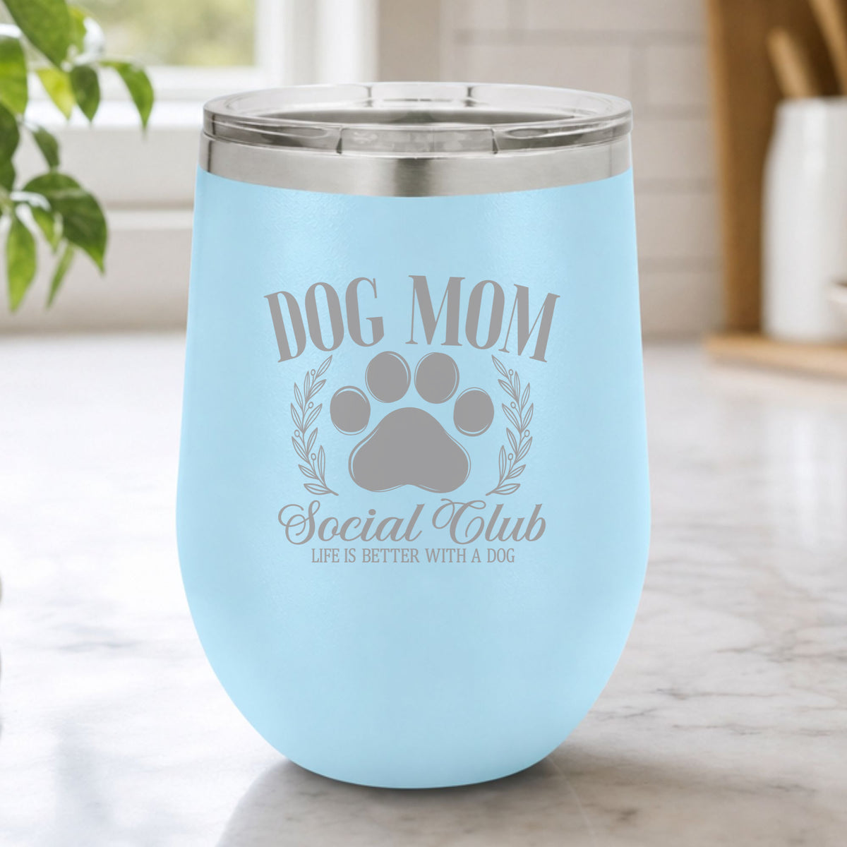 Light Blue 12 oz stemless wine tumbler laser engraved with the Social Club "Dog Mom" design, on a kitchen counter