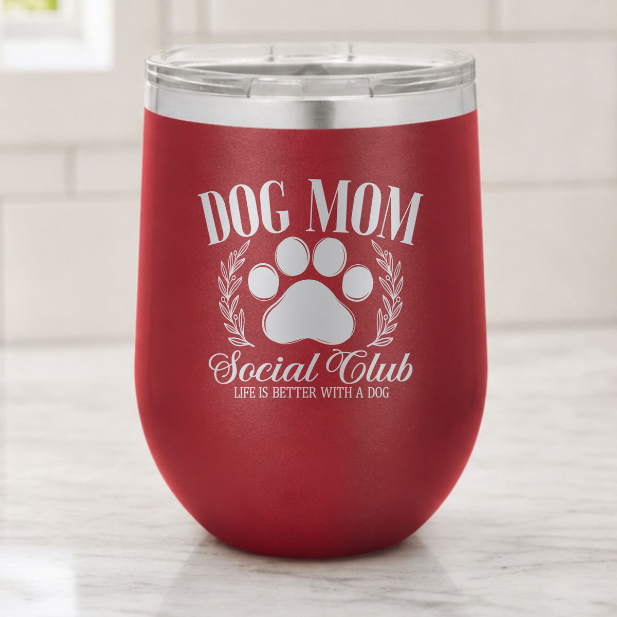 Maroon 12 oz stemless wine tumbler laser engraved with the Social Club "Dog Mom" design, on a kitchen counter