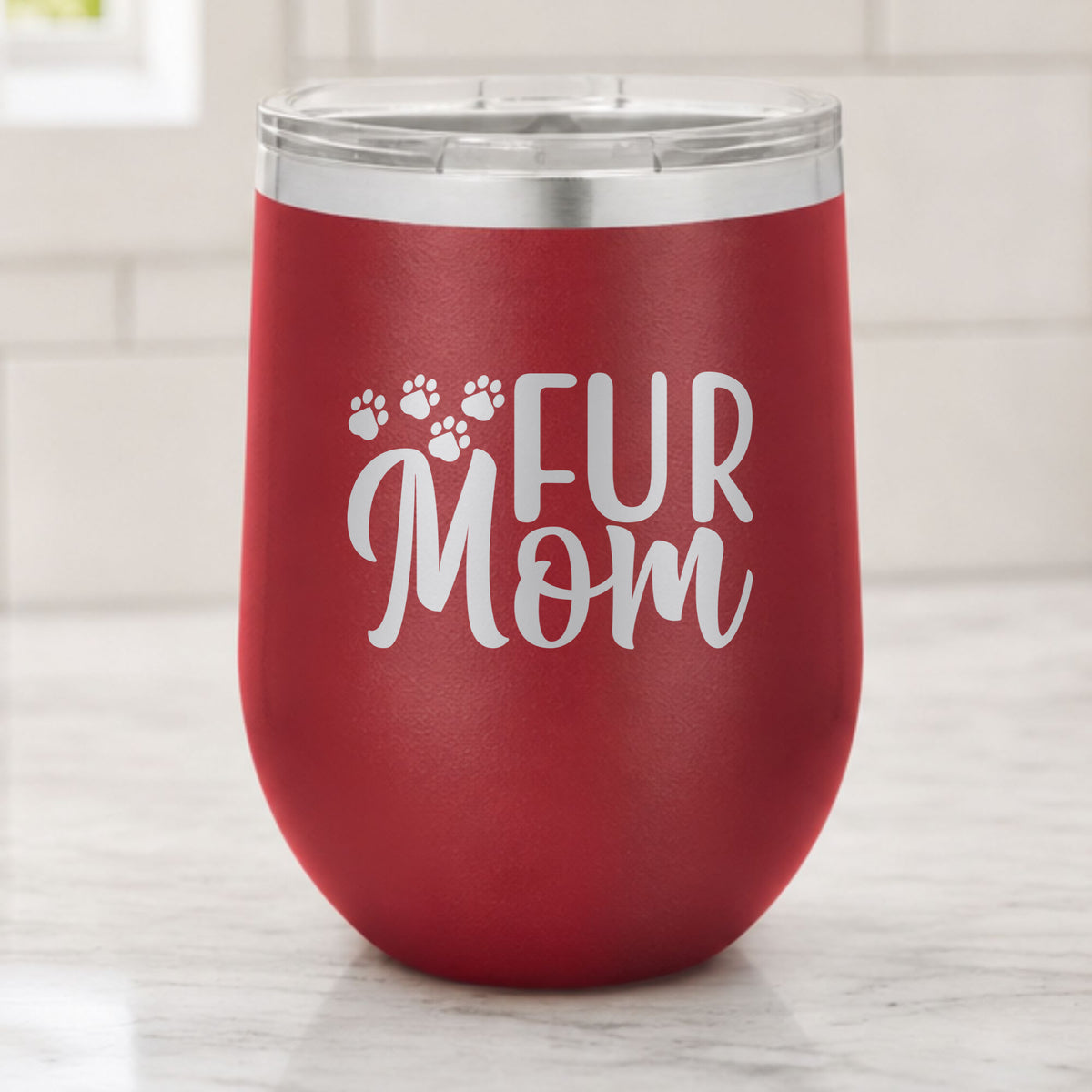 Maroon 12 oz stemless wine tumbler laser engraved with "Fur Mom", plus paw prints, on a kitchen counter