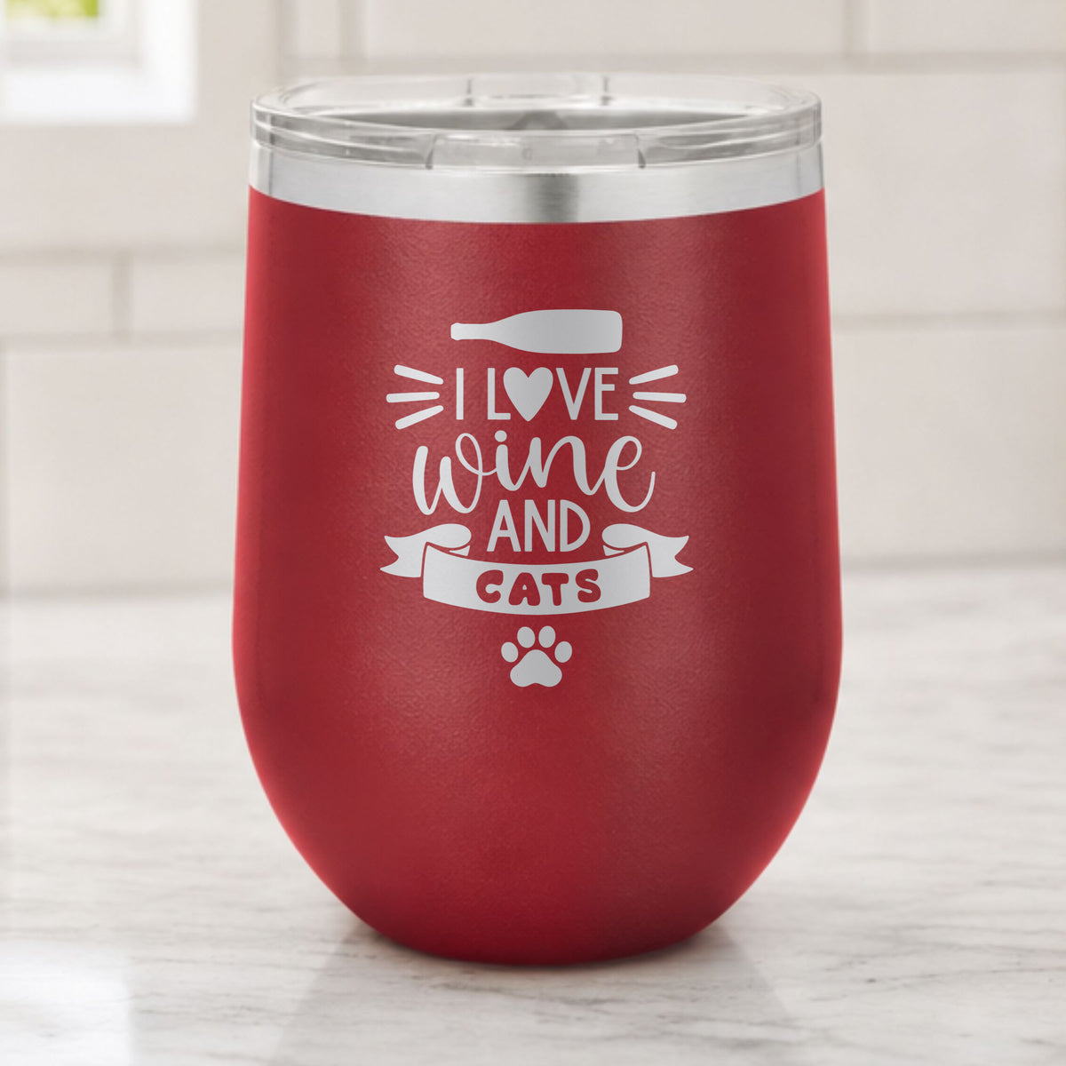 Maroon 12 oz stemless wine tumbler laser engraved with "I Love Wine and Cats", a wine bottle, a heart and a paw print, on a kitchen counter