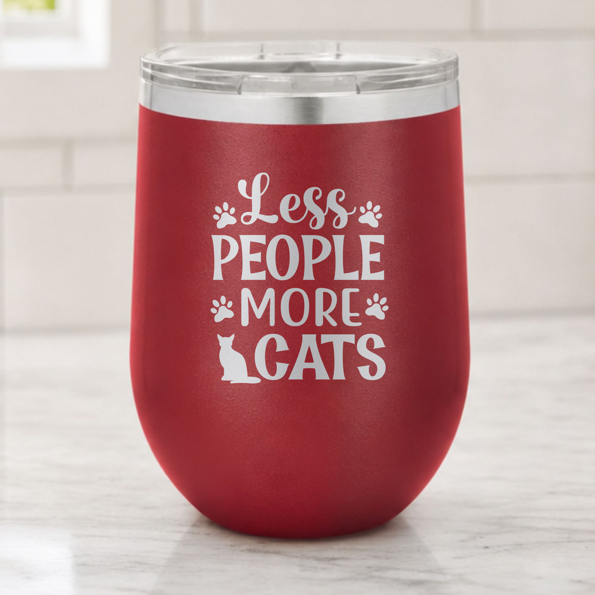 Maroon 12 oz stemless wine tumbler laser engraved with "Less People, More Cats", plus paw prints and a sitting cat, on a kitchen counter