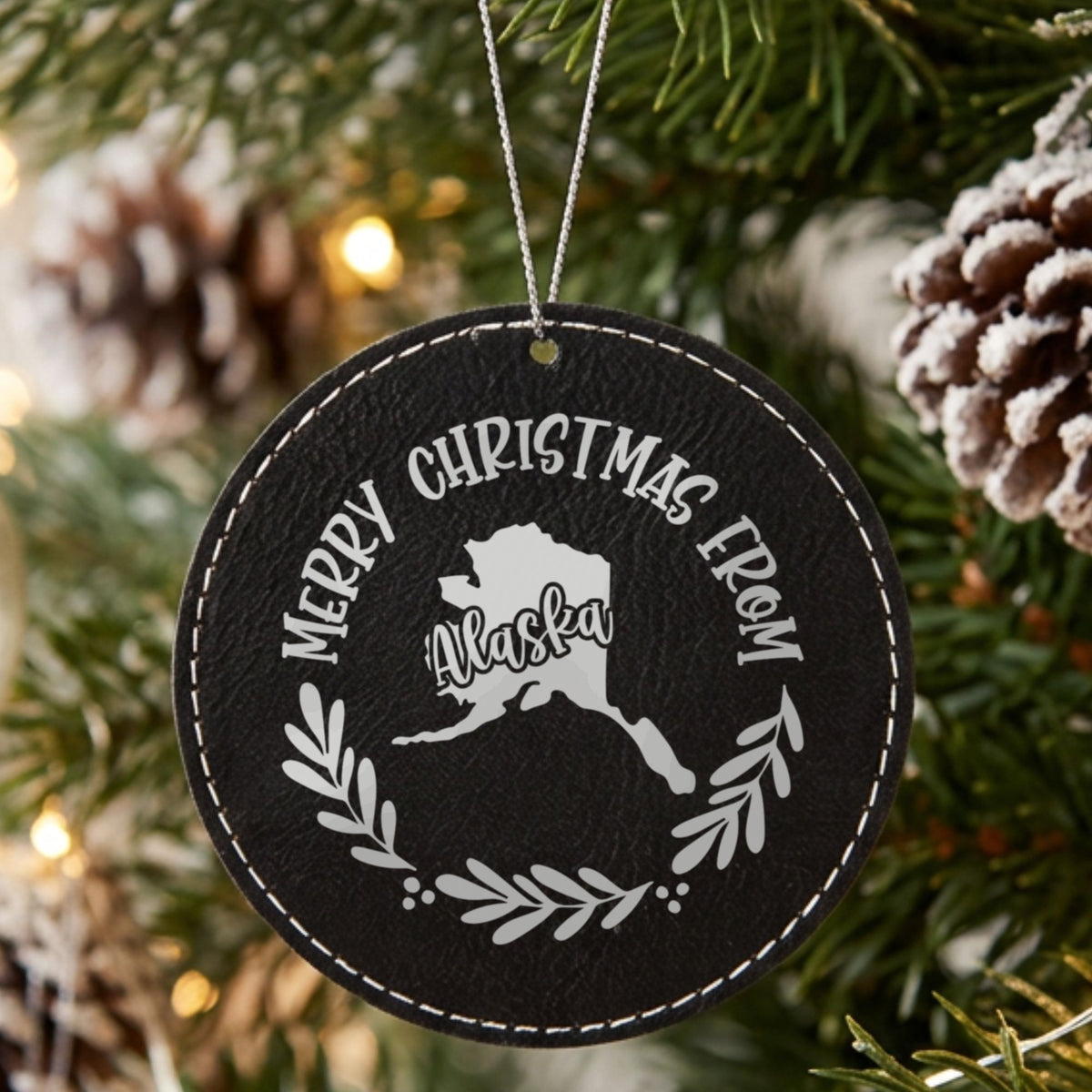 Black leatherette round Christmas ornament engraved Merry Christmas From Alaska, with the state shape in a laurel wreath, hanging on a tree