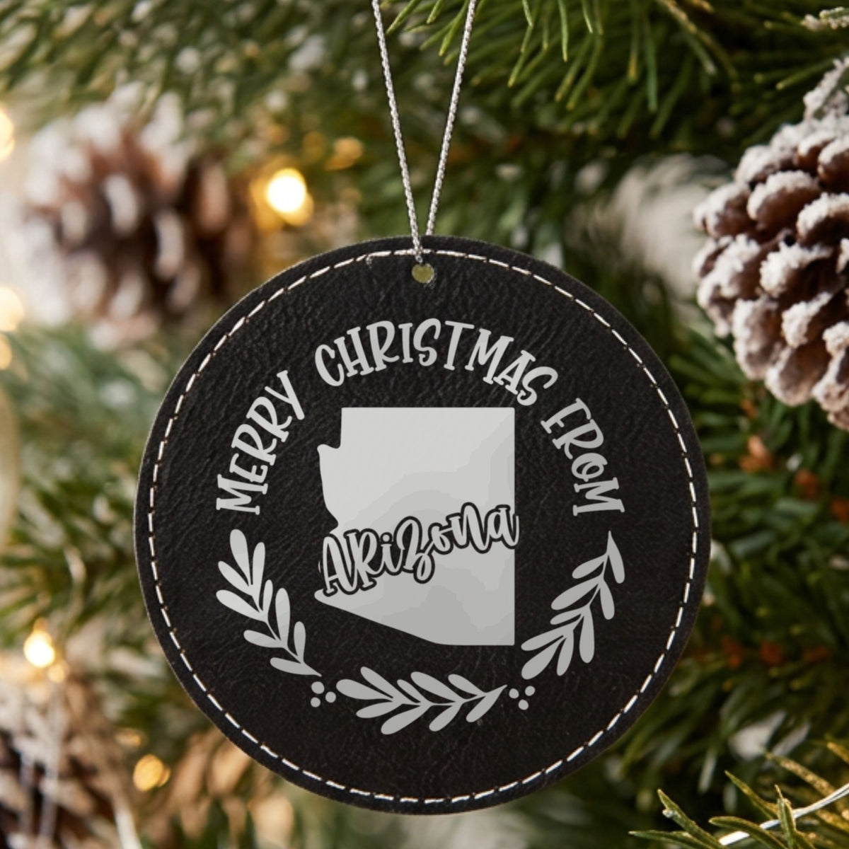 Black leatherette round Christmas ornament engraved Merry Christmas From Arizona, with the state shape in a laurel wreath, hanging on a tree