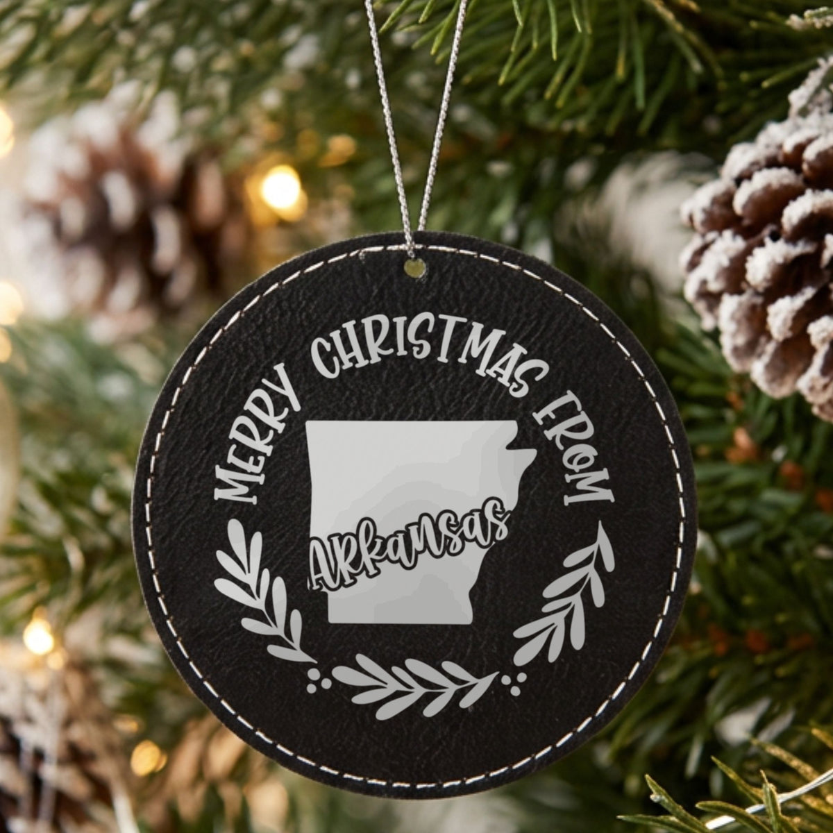 Black leatherette round Christmas ornament engraved Merry Christmas From Arkansas, with the state shape in a laurel wreath, hanging on a tree