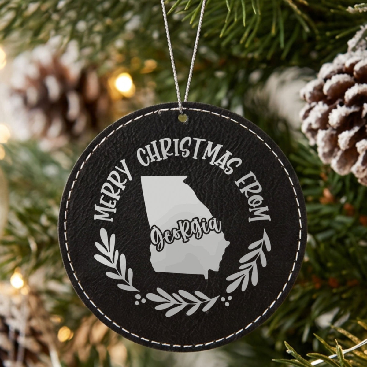 Black leatherette round Christmas ornament engraved Merry Christmas From Georgia, with the state shape in a laurel wreath, hanging on a tree