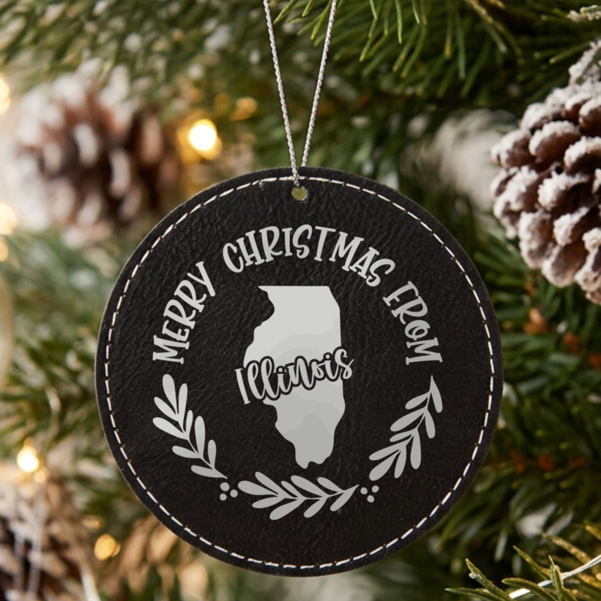 Black leatherette round Christmas ornament engraved Merry Christmas From Illinois, with the state shape in a laurel wreath, hanging on a tree