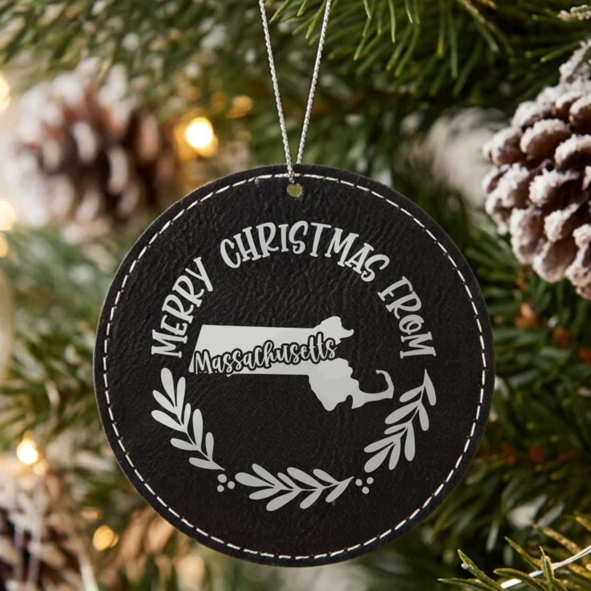 Black leatherette round Christmas ornament engraved Merry Christmas From Massachusetts, with the state shape in a laurel wreath, hanging on a tree