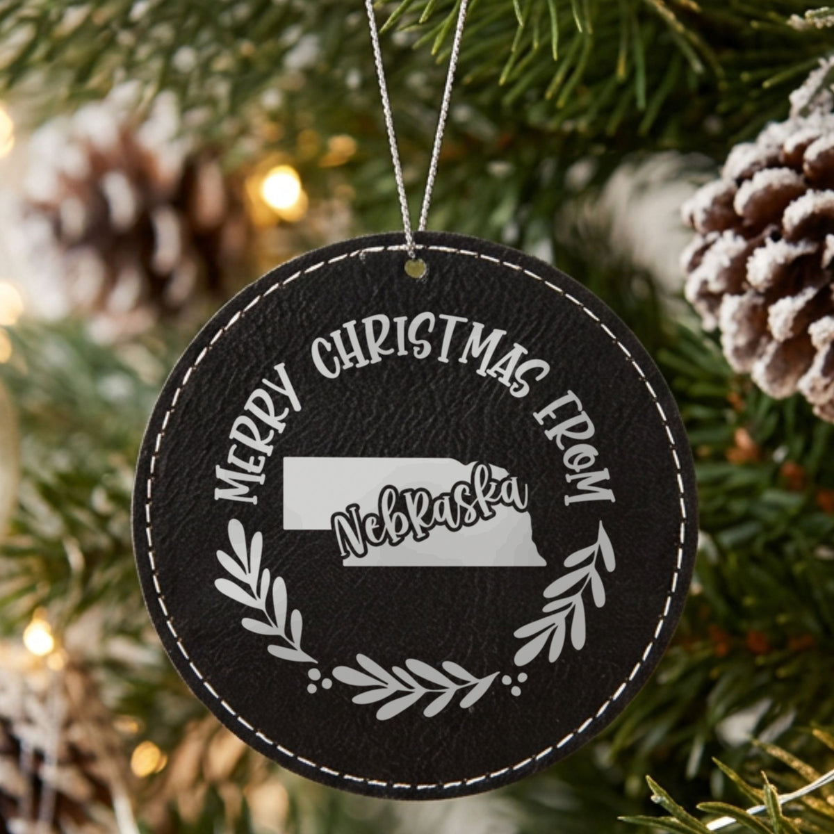 Black leatherette round Christmas ornament engraved Merry Christmas From Nebraska, with the state shape in a laurel wreath, hanging on a tree