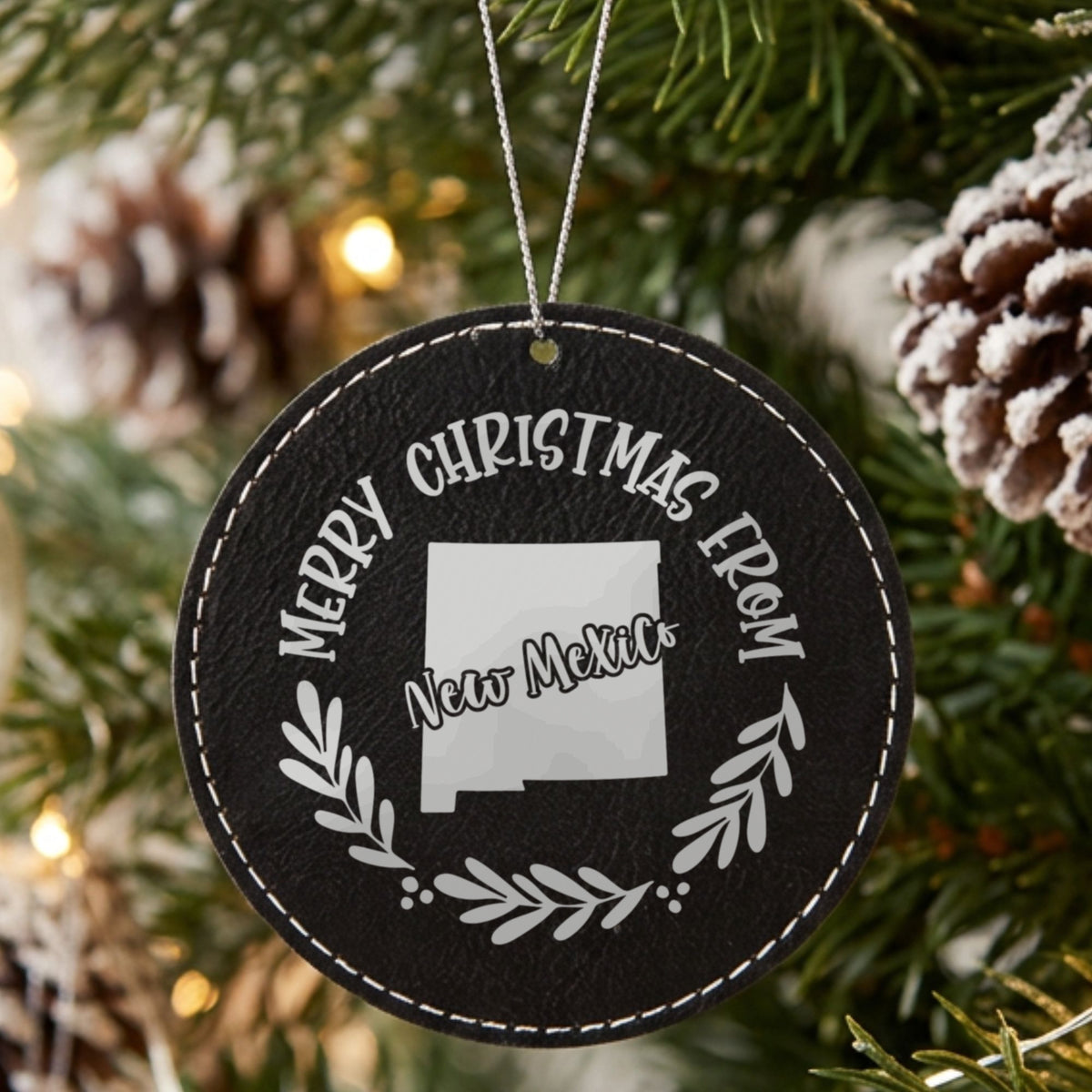 Black leatherette round Christmas ornament engraved Merry Christmas From New Mexico, with the state shape in a laurel wreath, hanging on a tree