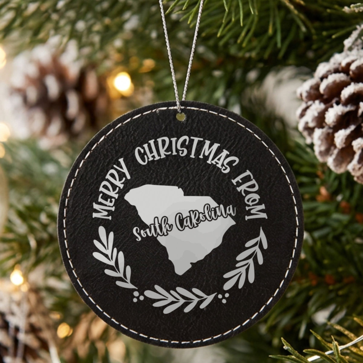 Black leatherette round Christmas ornament engraved Merry Christmas From South Carolina, with the state shape in a laurel wreath, hanging on a tree