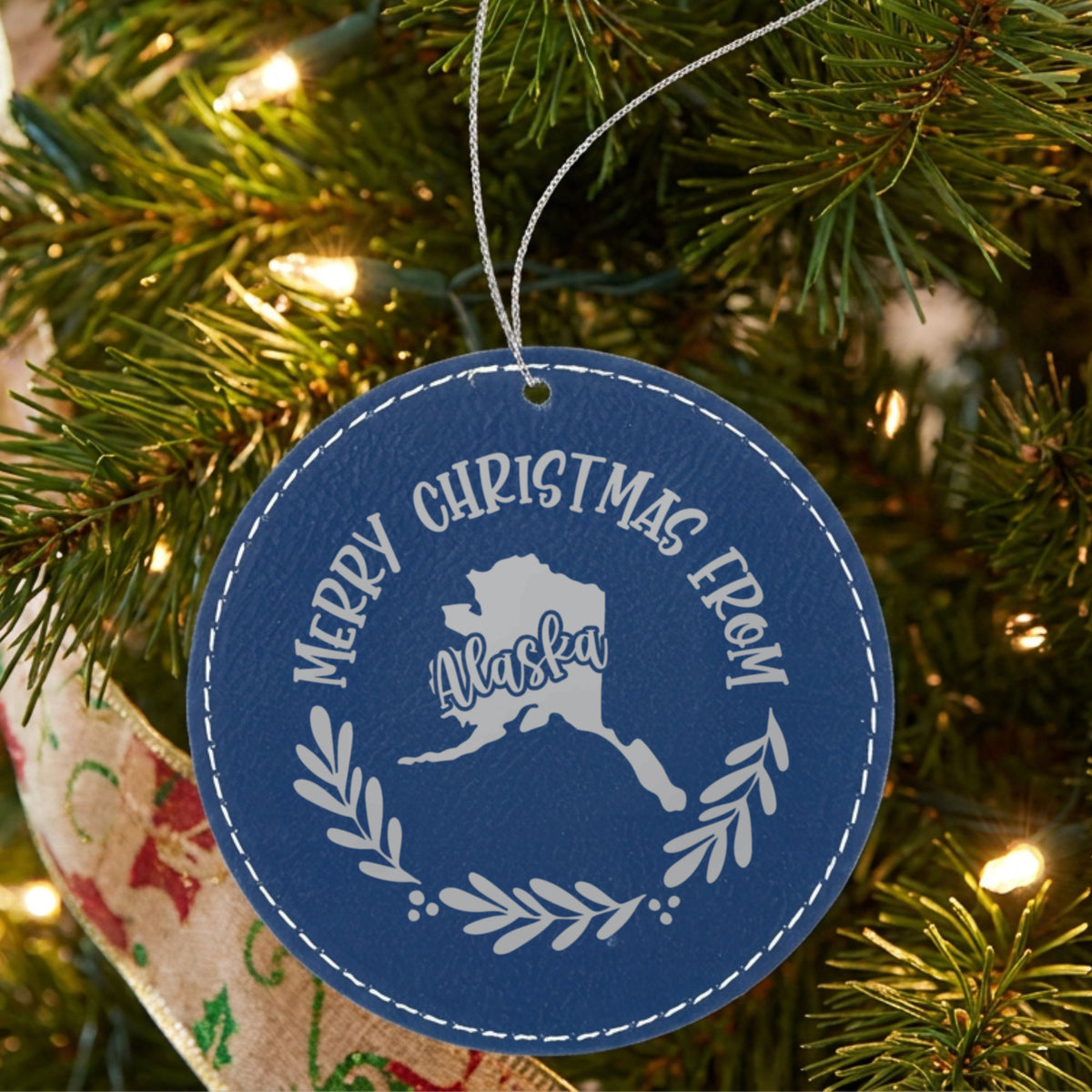 Blue leatherette round Christmas ornament engraved Merry Christmas From Alaska, with the state shape in a laurel wreath, hanging on a tree