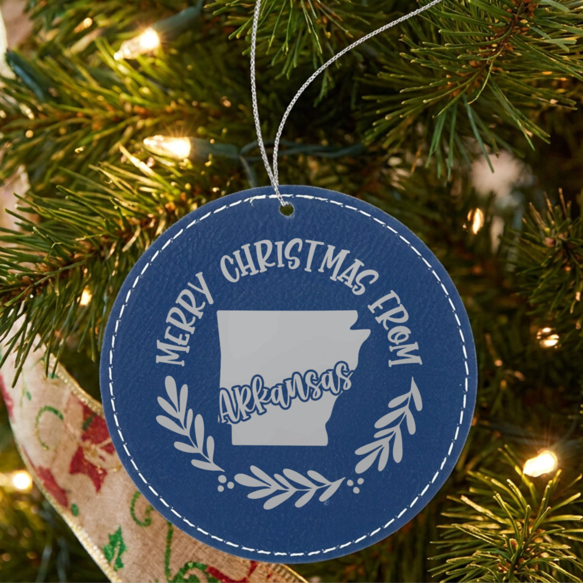 Blue leatherette round Christmas ornament engraved Merry Christmas From Arkansas, with the state shape in a laurel wreath, hanging on a tree