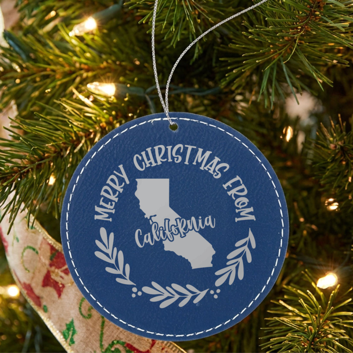 Blue leatherette round Christmas ornament engraved Merry Christmas From California, with the state shape in a laurel wreath, hanging on a tree