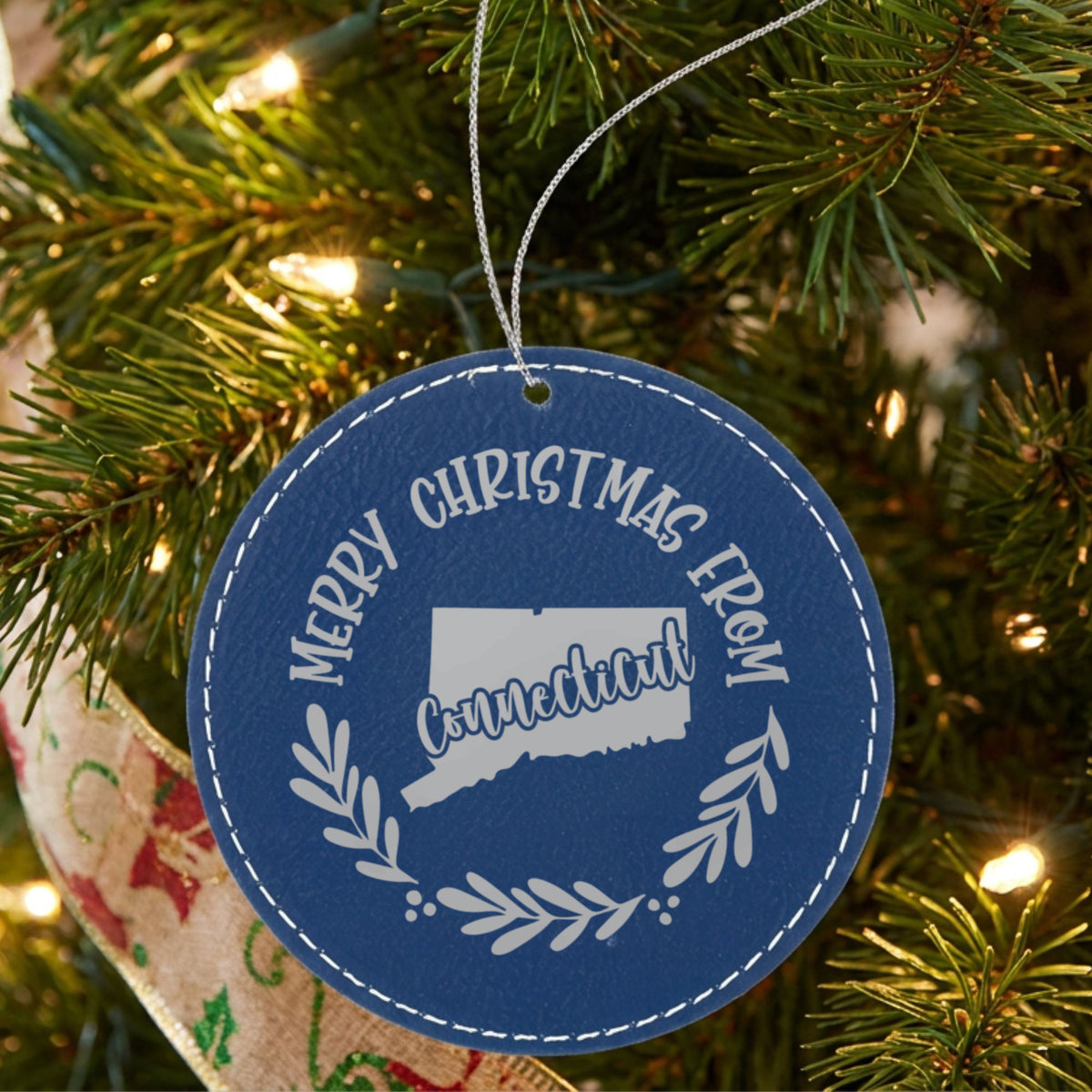 Blue leatherette round Christmas ornament engraved Merry Christmas From Connecticut, with the state shape in a laurel wreath, hanging on a tree