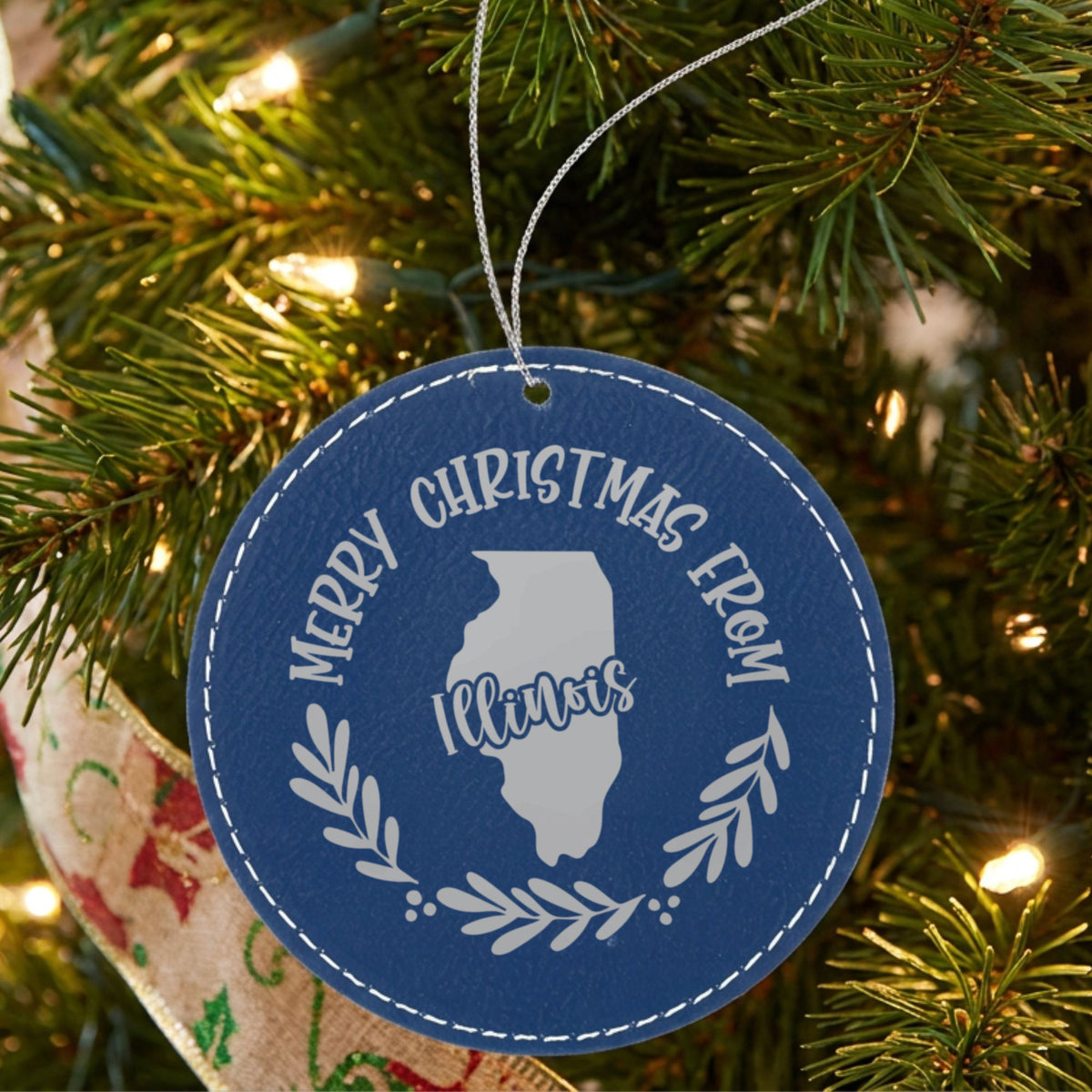 Blue leatherette round Christmas ornament engraved Merry Christmas From Illinois, with the state shape in a laurel wreath, hanging on a tree