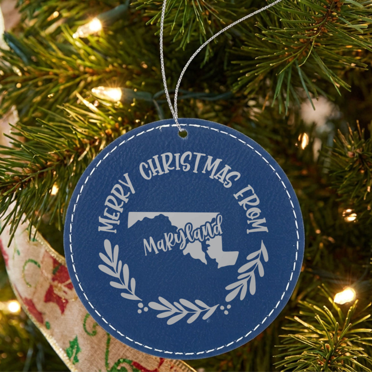 Blue leatherette round Christmas ornament engraved Merry Christmas From Maryland, with the state shape in a laurel wreath, hanging on a tree