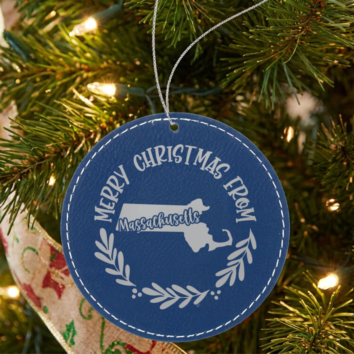 Blue leatherette round Christmas ornament engraved Merry Christmas From Massachusetts, with the state shape in a laurel wreath, hanging on a tree