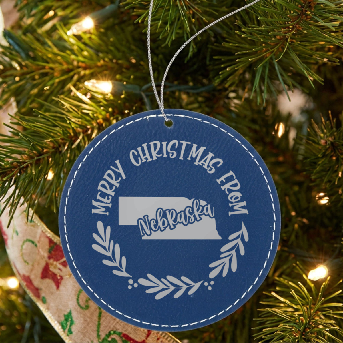 Blue leatherette round Christmas ornament engraved Merry Christmas From Nebraska, with the state shape in a laurel wreath, hanging on a tree