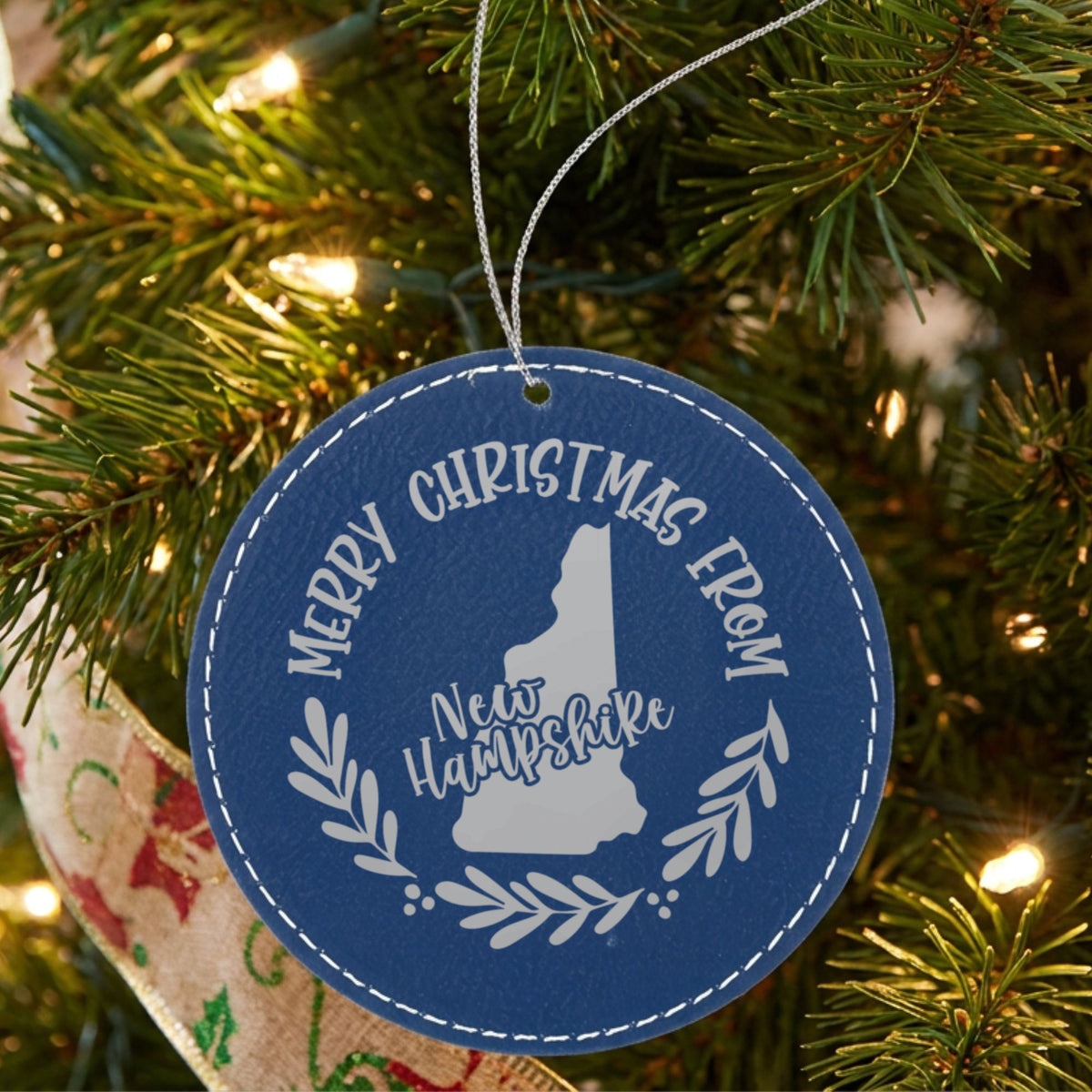 Blue leatherette round Christmas ornament engraved Merry Christmas From New Hampshire, with the state shape in a laurel wreath, hanging on a tree