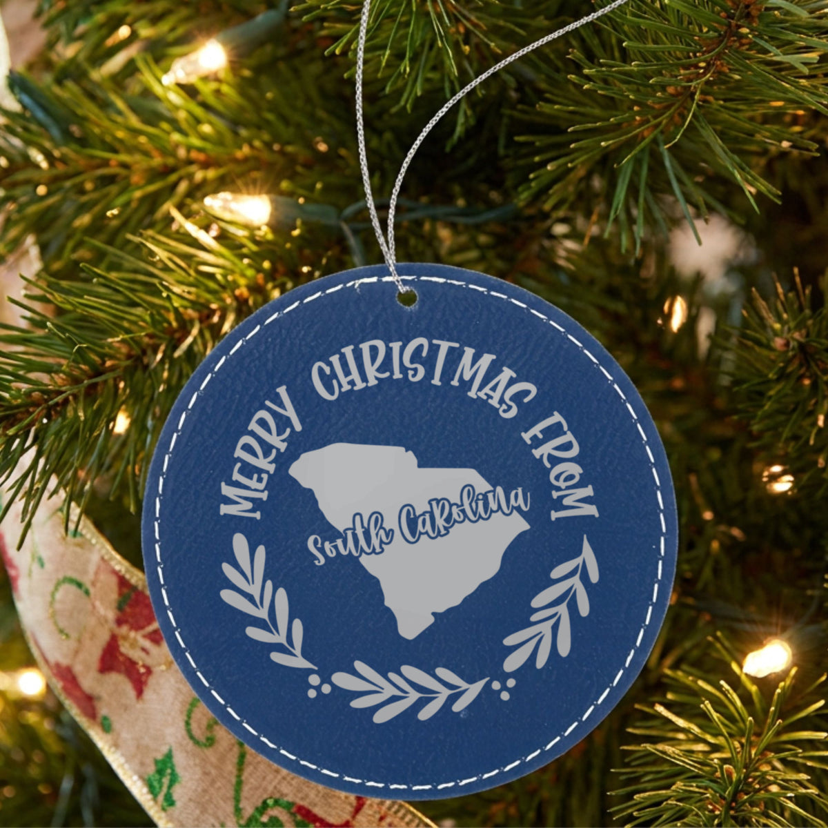 Blue leatherette round Christmas ornament engraved Merry Christmas From South Carolina, with the state shape in a laurel wreath, hanging on a tree