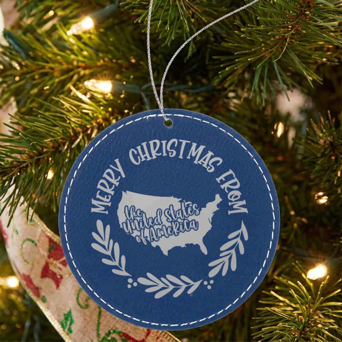 Blue leatherette round Christmas ornament engraved Merry Christmas From the USA, with the country shape in a laurel wreath, hanging on a tree
