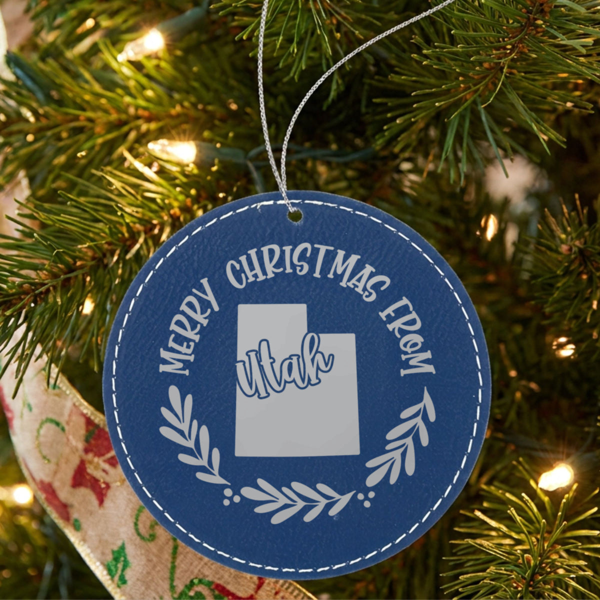 Blue leatherette round Christmas ornament engraved Merry Christmas From Utah, with the state shape in a laurel wreath, hanging on a tree