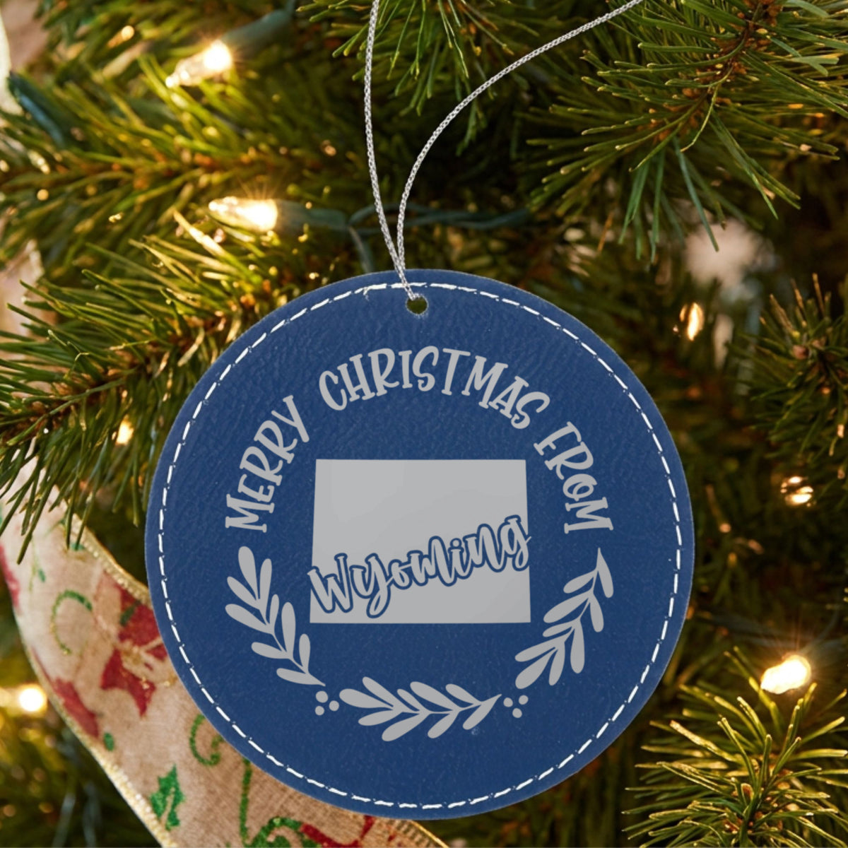 Blue leatherette round Christmas ornament engraved Merry Christmas From Wyoming, with the state shape in a laurel wreath, hanging on a tree