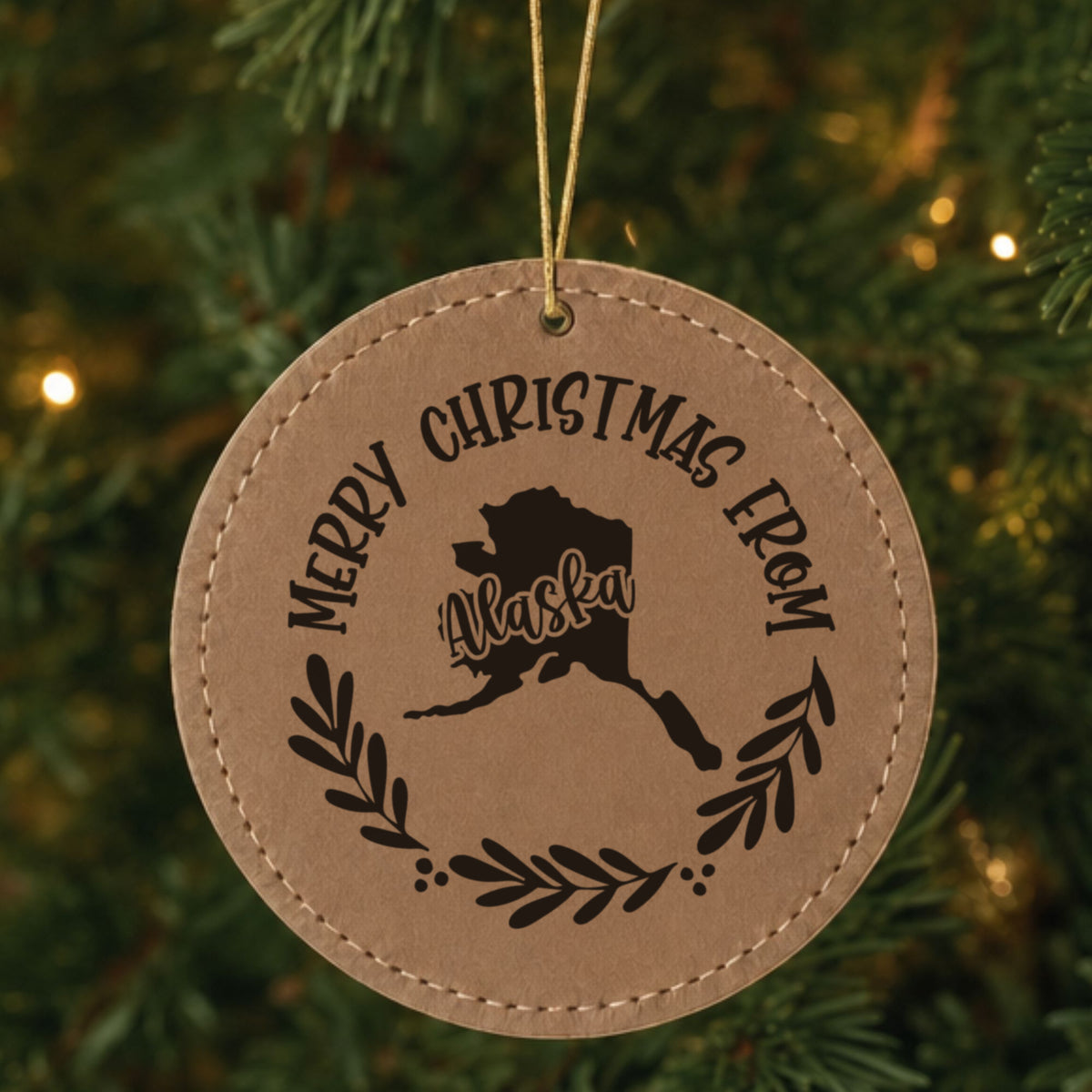 Brown leatherette round Christmas ornament engraved Merry Christmas From Alaska, with the state shape in a laurel wreath, hanging on a tree
