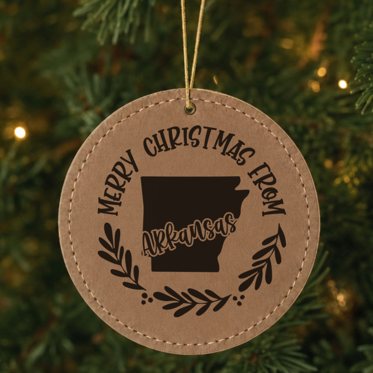 Brown leatherette round Christmas ornament engraved Merry Christmas From Arkansas, with the state shape in a laurel wreath, hanging on a tree
