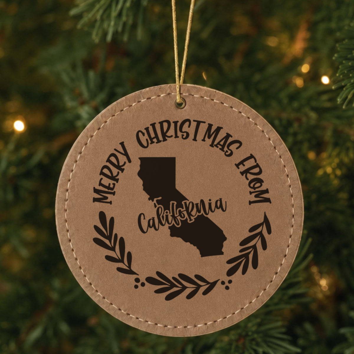 Brown leatherette round Christmas ornament engraved Merry Christmas From California, with the state shape in a laurel wreath, hanging on a tree