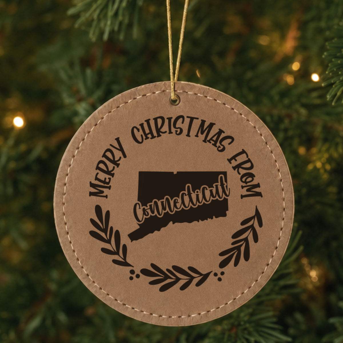 Brown leatherette round Christmas ornament engraved Merry Christmas From Connecticut, with the state shape in a laurel wreath, hanging on a tree