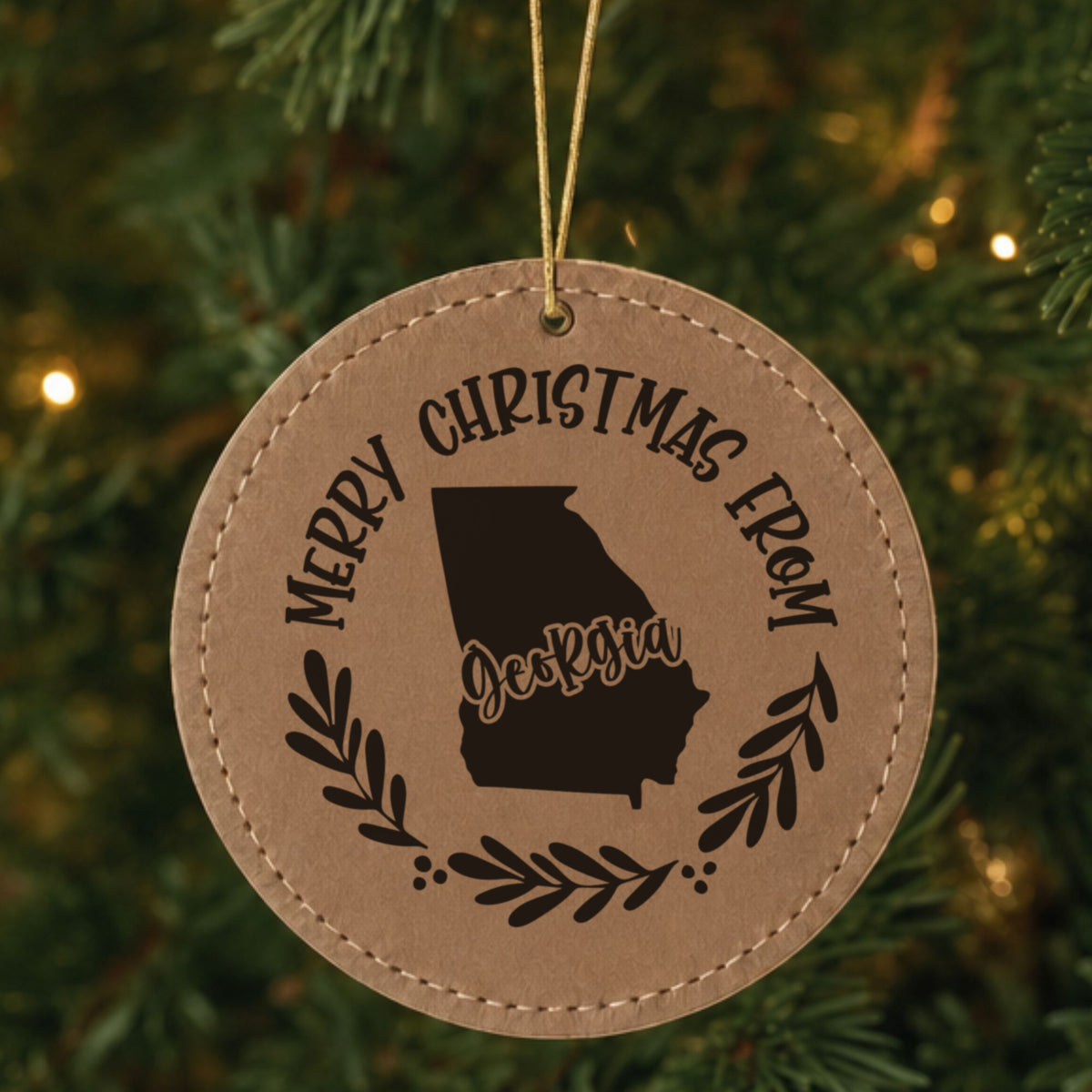 Brown leatherette round Christmas ornament engraved Merry Christmas From Georgia, with the state shape in a laurel wreath, hanging on a tree