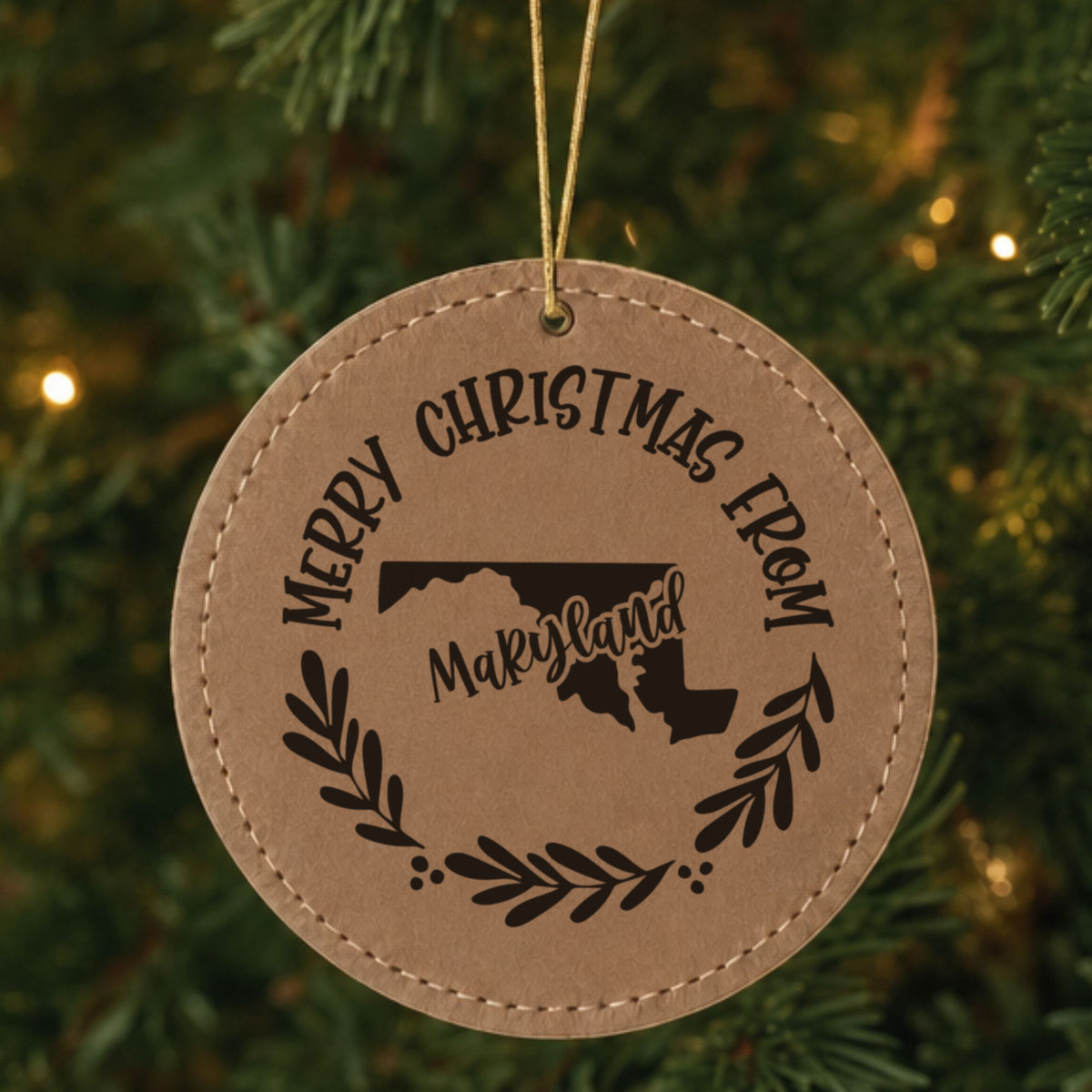 Brown leatherette round Christmas ornament engraved Merry Christmas From Maryland, with the state shape in a laurel wreath, hanging on a tree