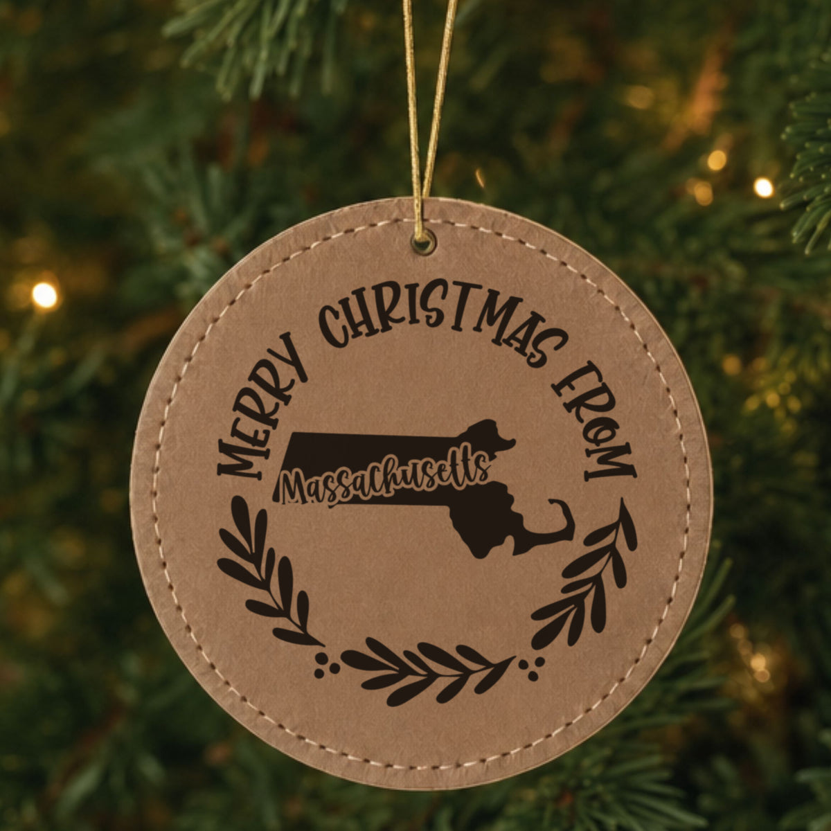 Brown leatherette round Christmas ornament engraved Merry Christmas From Massachusetts, with the state shape in a laurel wreath, hanging on a tree