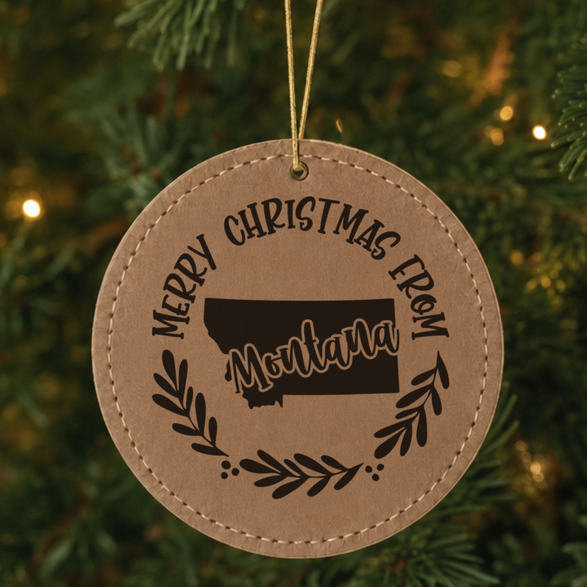 Brown leatherette round Christmas ornament engraved Merry Christmas From Montana, with the state shape in a laurel wreath, hanging on a tree