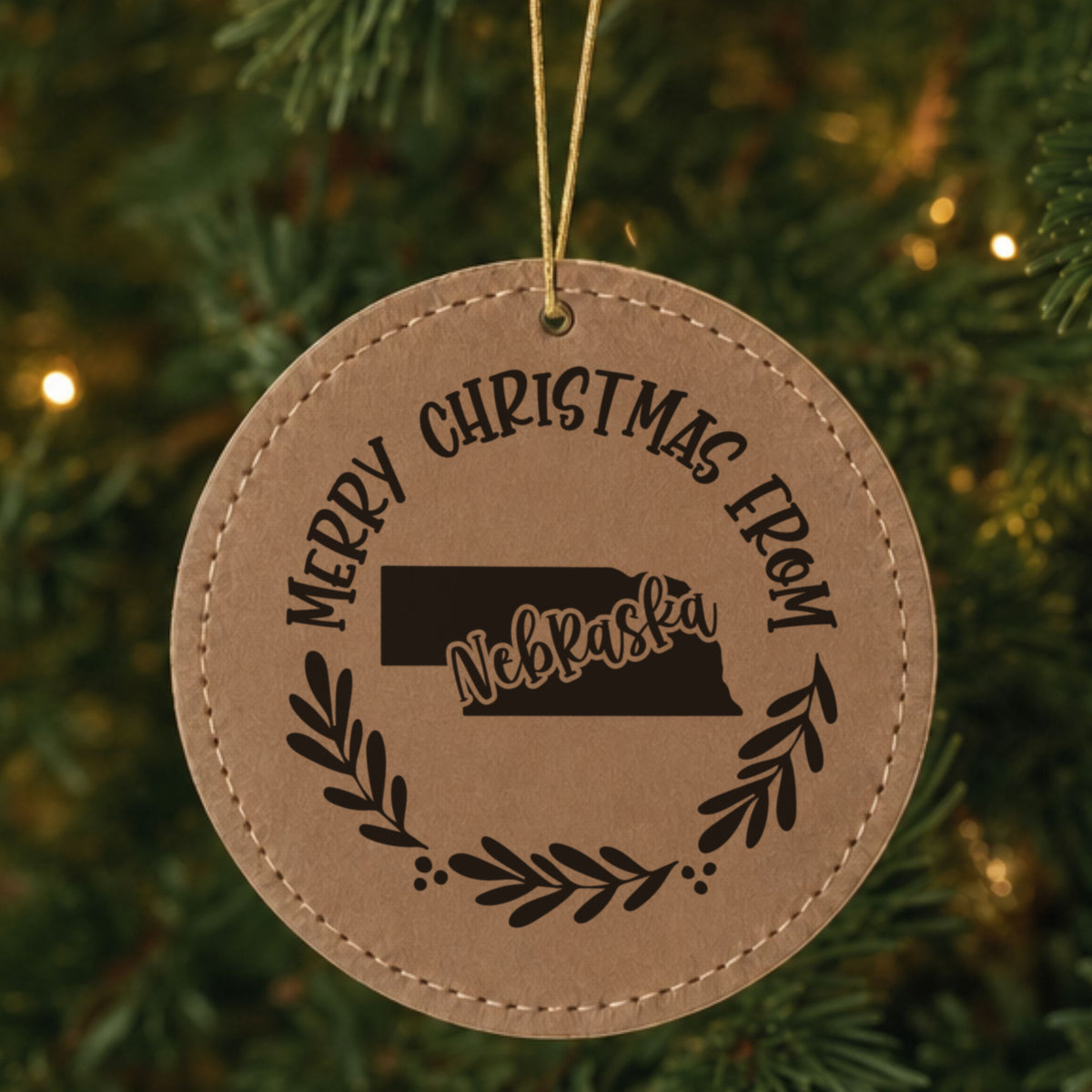 Brown leatherette round Christmas ornament engraved Merry Christmas From Nebraska, with the state shape in a laurel wreath, hanging on a tree