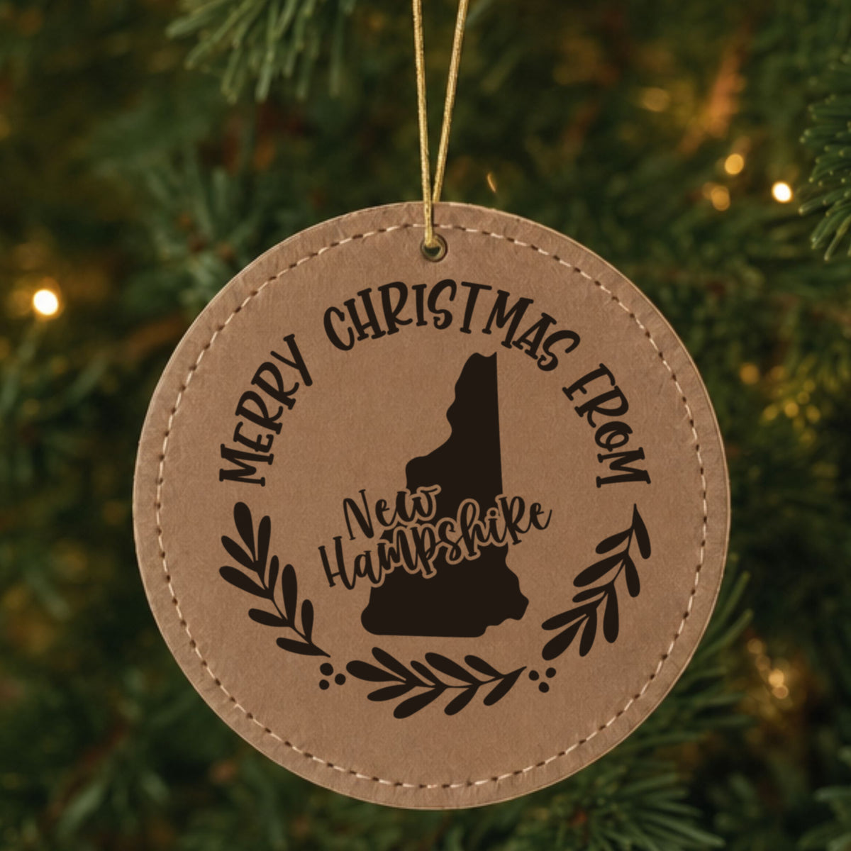 Brown leatherette round Christmas ornament engraved Merry Christmas From New Hampshire, with the state shape in a laurel wreath, hanging on a tree