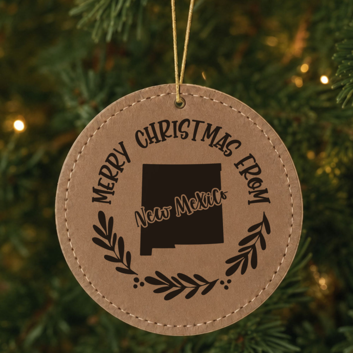 Brown leatherette round Christmas ornament engraved Merry Christmas From New Mexico, with the state shape in a laurel wreath, hanging on a tree