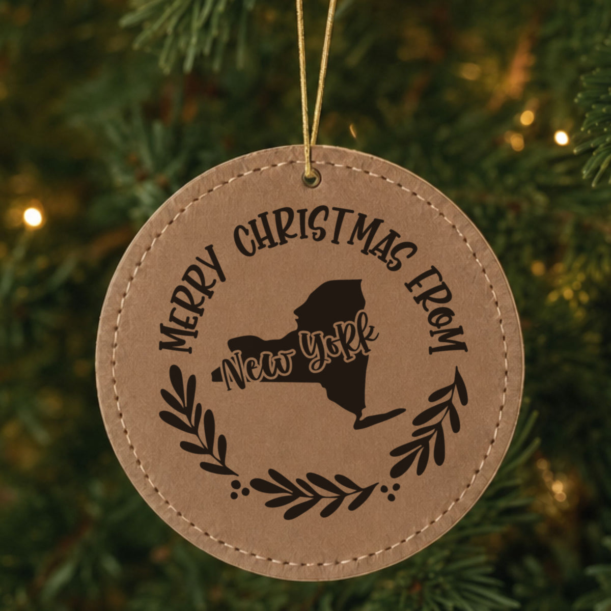 Brown leatherette round Christmas ornament engraved Merry Christmas From New York, with the state shape in a laurel wreath, hanging on a tree