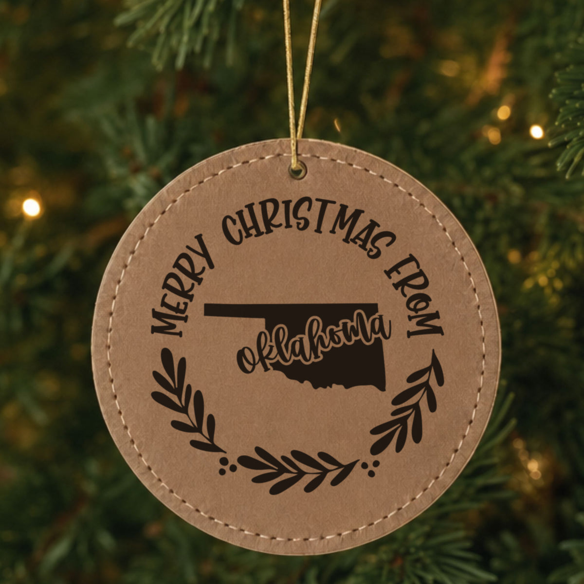 Brown leatherette round Christmas ornament engraved Merry Christmas From Oklahoma, with the state shape in a laurel wreath, hanging on a tree